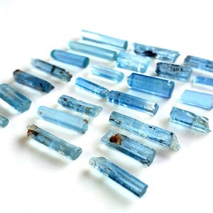 May include: A collection of twenty-four raw, elongated aquamarine crystal specimens arranged in a grid. These natural gemstones exhibit varying shades of translucent light blue, with some pieces showing internal inclusions and rough, textured surfaces.