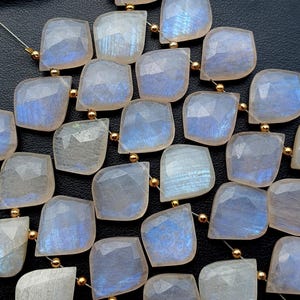 May include: A collection of moonstone beads, each bead is a square shape with rounded corners, showing a mix of pale blue and white tones. The beads are strung together with gold-coloured spacers, creating a shimmering effect. The background is a dark, solid colour.
