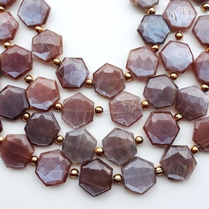 May include: A collection of faceted hexagonal beads in shades of dusty mauve and muted purple, arranged on thin wire strands. Small, round, metallic gold-toned spacer beads are interspersed between the larger hexagonal stone beads throughout.