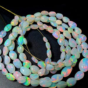 Puede incluir: Cadenas de cuentas de ópalo ovaladas e iridiscentes. Los ópalos muestran un color base blanco lechoso con destellos de rojo, verde y azul. Las cuentas están ensartadas en un fino alambre dorado, ideal para joyería.