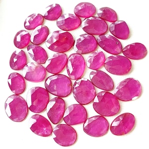 Pode incluir: Uma coleção de pedras preciosas de rubi facetadas, em forma oval. As gemas, de cor vermelho-rosada vibrante, variam ligeiramente em tamanho. Estão dispostas sobre uma superfície branca, mostrando suas superfícies polidas.