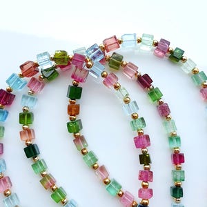 Op de afbeelding: Close-up van verschillende strengen kleurrijke edelsteenkettingen. De kettingen hebben kleine, vierkant geslepen edelstenen in verschillende kleuren, waaronder roze, groen, blauw en oranje, afgewisseld met kleine gouden kralen. De edelstenen zijn aan elkaar geregen, waardoor een levendig ontwerp ontstaat.
