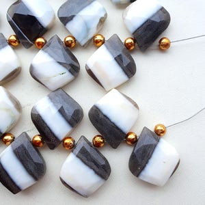 May include: Close-up of a strand of gemstone beads. Each bead is a faceted, diamond-shaped stone with a diagonal stripe of white and dark gray. Small gold-colored beads separate the larger stones. The beads are strung on a thin, clear cord.