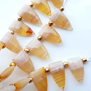 May include: A strand of faceted, triangular-shaped yellow agate beads with translucent, banded patterns. Each stone is separated by a small, round, metallic gold-colored spacer bead, strung together on a thin, clear filament wire.
