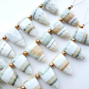May include: A collection of faceted, triangular-shaped stone beads arranged in rows. Each bead features horizontal bands of white, beige, and pale green, with some showing dark speckles. Small, round, metallic gold-colored spacer beads separate them.