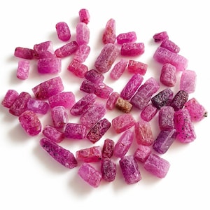 May include: A collection of small, raw, cylindrical ruby gemstones in various shades of vibrant pink and deep magenta. Each stone features a rough, textured surface with natural crystalline facets and irregular edges, scattered loosely across the frame.