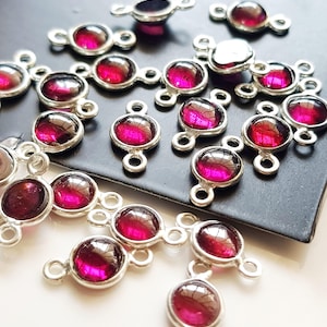 May include: A collection of silver-toned metal jewelry connectors, each featuring a circular, deep magenta-colored cabochon stone. These small components have two loops, one on each side, designed for linking chains or other jewelry elements together.