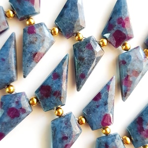 Könnte beinhalten: Ein Strang aus facettierten, trapezförmigen blauen Kyanit-Perlen mit eingebetteten magentafarbenen Rubineinschlüssen, durchsetzt mit kleinen, runden, polierten goldfarbenen Metall-Zwischenperlen auf einem dünnen Faden.