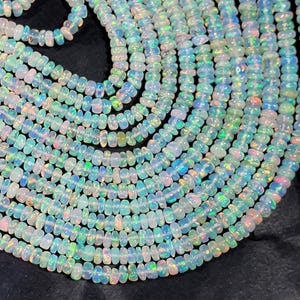 May include: A collection of multiple strands of small, round, translucent opal beads. Each bead displays a milky white base color with iridescent flashes of green, blue, pink, and orange light, arranged in parallel rows on a dark surface.