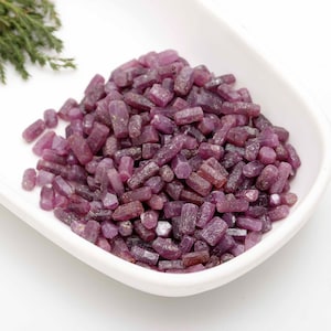 May include: A white, shallow dish filled with a pile of raw, uncut ruby gemstones. The gemstones are a deep reddish-purple color and have a rough, natural texture. A sprig of green foliage is in the upper left corner.
