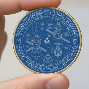 May include: A round, gold-rimmed coin featuring a blue blueprint design. The blueprint includes technical diagrams and text related to the Artemis II Orion spacecraft. The coin is held in a hand.