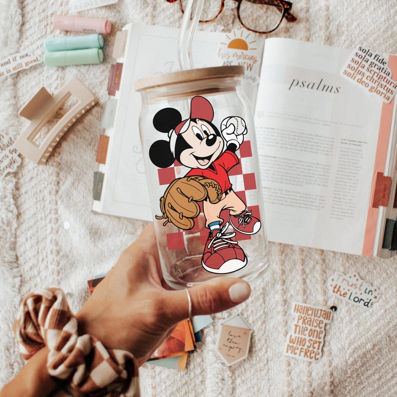 May include: Clear glass tumbler with a wooden lid and straw. The tumbler features a baseball-themed Mickey Mouse graphic. The design includes a red and white checkered pattern. The tumbler is held by a person.
