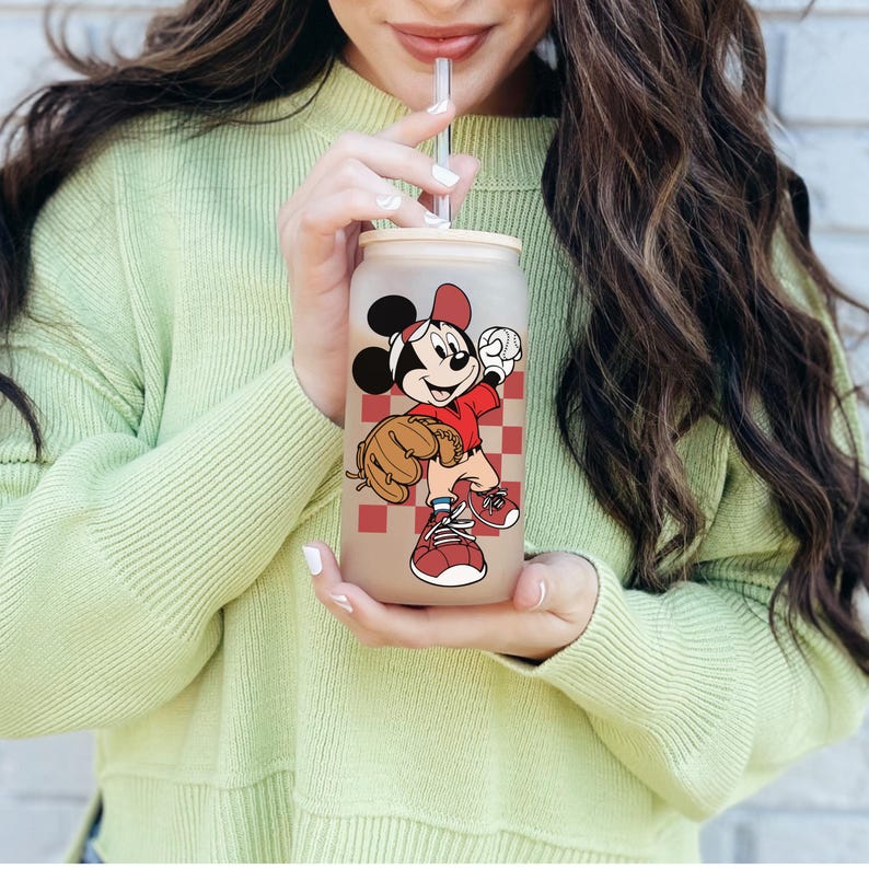 May include: A frosted glass tumbler with a wooden lid and a straw. The tumbler features a cartoon image of Mickey Mouse in a baseball uniform, set against a red and white checkered background. The design includes a baseball glove and a baseball.