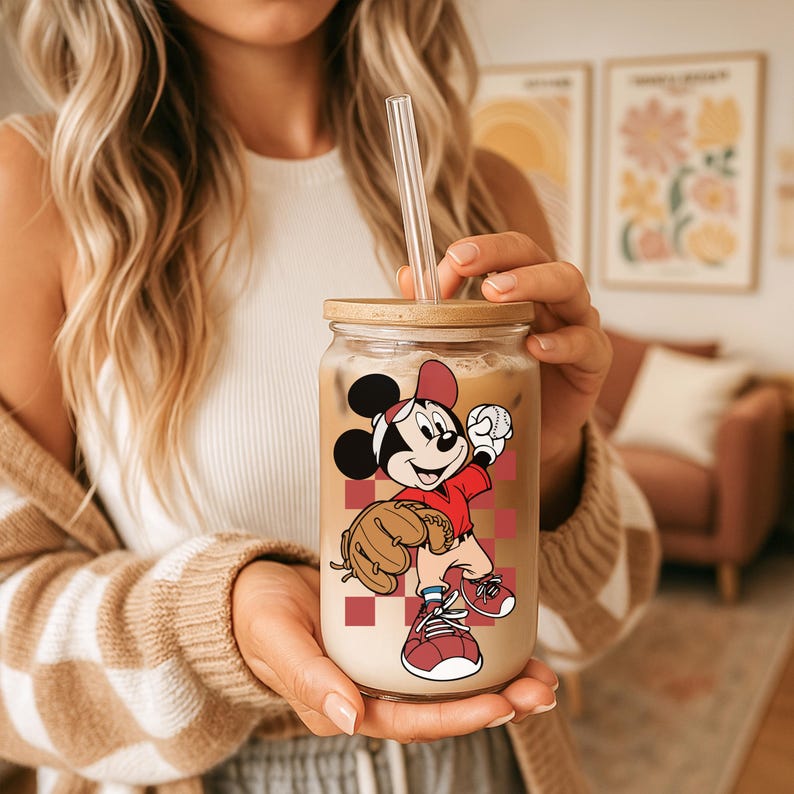 May include: A clear glass tumbler with a bamboo lid and a straw, featuring a cartoon image of Mickey Mouse in a baseball uniform. The tumbler is filled with a creamy beverage. The design includes a baseball glove and a red and white checkered background.