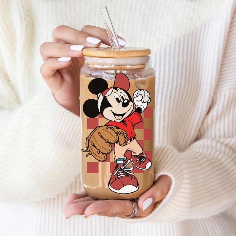 May include: A clear glass tumbler with a bamboo lid and a clear straw. The tumbler features a cartoon image of Mickey Mouse in a baseball uniform, holding a baseball and wearing a glove. The background has a red and brown checkered pattern.