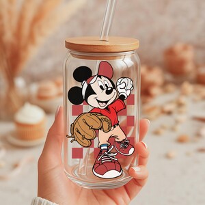 May include: A clear glass tumbler with a wooden lid and a straw. The tumbler features a cartoon illustration of Mickey Mouse in a baseball uniform, holding a baseball and a glove. The background has a red and white checkered pattern.