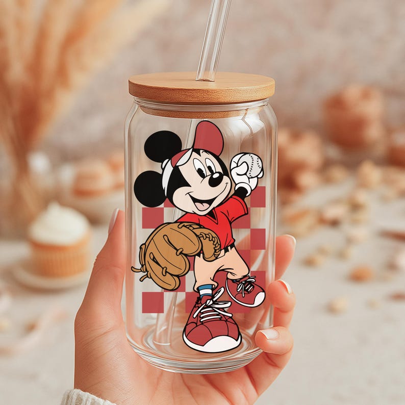 May include: A clear glass tumbler with a wooden lid and a straw. The tumbler features a cartoon illustration of Mickey Mouse in a baseball uniform, holding a baseball and a glove. The background has a red and white checkered pattern.