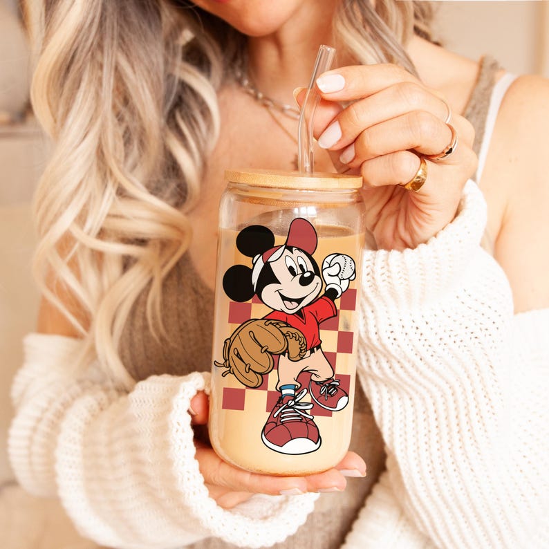 May include: A clear glass tumbler with a bamboo lid and a straw. The tumbler features a cartoon image of Mickey Mouse in a baseball uniform, holding a baseball and glove. The background has a red and brown checkered pattern.