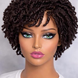May include: A close-up of a mannequin head with a short, dark brown curly wig. The mannequin has green eyes, dark eyeliner, and pink lipstick. The mannequin is wearing a white t-shirt.