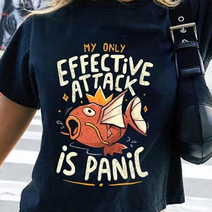 May include: Black crop top t-shirt featuring the text "MY ONLY EFFECTIVE ATTACK IS PANIC" and a cartoon fish design in orange, yellow, and white. The shirt has short sleeves and a round neckline.