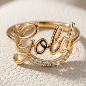 May include: A gold ring with the word "Gold" in cursive script. The ring features a band of small, clear gemstones. The ring is set against a neutral background.