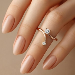 Op de afbeelding: Een roségouden ring met twee heldere, ronde edelstenen. Een grotere edelsteen is aan de bovenkant gezet, de andere kleiner eronder. De ring heeft een uniek, gebogen ontwerp.