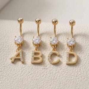 Op de afbeelding: Goudkleurige navelringen met de letters A, B, C en D. Elke ring heeft een sprankelende, heldere edelsteen en een bungelende initiaal. De letters zijn versierd met kleine, heldere stenen.