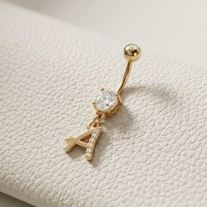 May include: Gold-coloured belly button ring with a dangling letter "A" embellished with small clear stones. The ring also features a large, round, clear stone and a small clear stone on the top ball.