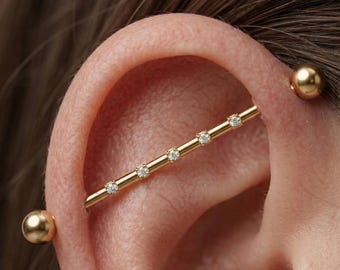 May include: A gold industrial barbell piercing jewelry featuring a straight bar with five evenly spaced small clear round-cut stones set along the center and two smooth polished gold spherical end balls securing the piece in the ear cartilage.