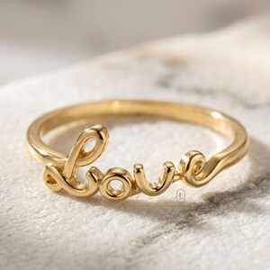 Può includere: Un anello color oro con la parola "love" in corsivo. L'anello è realizzato in metallo lucido e si trova su una superficie bianca. Questo delicato gioiello è un anello.