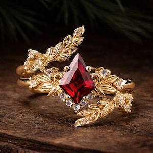 May include: A gold-colored ring with a large, red, diamond-shaped gemstone as the focal point. The ring features intricate leaf and floral designs, with smaller clear gemstones accenting the main stone. The ring is set on a wooden surface.