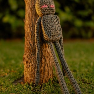 May include: A handmade grey crochet doll with long limbs and a rounded head, leaning against a tree. The doll has pink accents on its face and is positioned on a grassy surface, suggesting a whimsical, handcrafted toy.