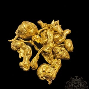 May include: A collection of irregularly shaped, golden-yellow gold nuggets against a black background. The nuggets vary in size and texture, with some appearing smooth and others more porous. The image includes the Noble Ingots logo in the bottom right corner.
