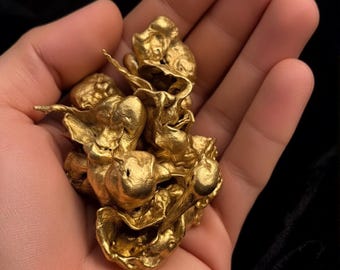 May include: A cluster of raw, metallic gold nuggets with an irregular, textured surface. The gold has a bright, reflective yellow finish. In the upper left corner, a circular logo features the text NOBLE INGOTS GOLD RECOVERY & MINTING.