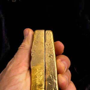 Può includere: Due lingotti d'oro rettangolari con superfici ruvide e strutturate sono tenuti in una mano. L'oro ha un aspetto brillante e lucente. Il logo "NOBLE INGOTS GOLD RECOVERY & MINTING" è visibile sullo sfondo.