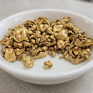 May include: A white ceramic bowl containing gold nuggets. The nuggets are of different sizes and shapes, with a bright, metallic gold colour. The bowl is round with a slightly raised edge.