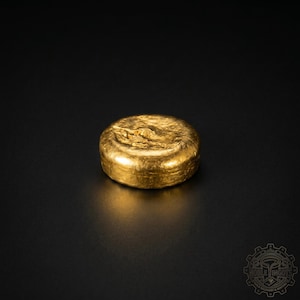 May include: A solid, round gold ingot with a textured surface. The gold bar is a bright, shiny yellow color and sits on a black surface. The ingot is a precious metal and is likely used for investment or collection.