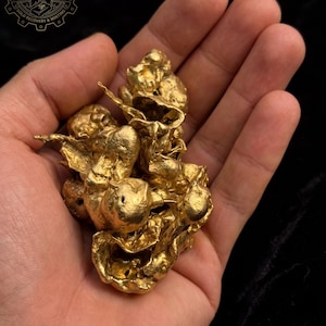 May include: A cluster of gold nuggets, displaying a bright, metallic yellow colour. The uneven shapes and textures of the gold are visible, set against a dark backdrop. The image includes the text "NOBLE INGOTS GOLD RECOVERY & MINTING".