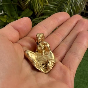 May include: A gold nugget with a rough, irregular shape. The nugget is held in an open hand, with a green and leafy background. The gold nugget appears to be solid and has a bright, reflective surface.