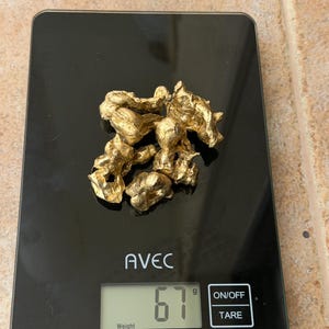 May include: A black digital scale shows "AVEC" and the weight of gold nuggets. The scale reads 67 grams. The nuggets are a bright, metallic gold colour and have a rough, irregular texture. The scale has an on/off and tare button.