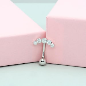 Op de afbeelding: Een zilverkleurige navelpiercing met een gebogen bovenkant versierd met vijf heldere, ronde edelstenen. De onderkant van de ring heeft een kleine, ronde bal. De sieraden zijn geplaatst tegen een zachtroze achtergrond.