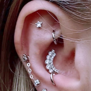 May include: Close-up of an ear with multiple piercings. The piercings include a small star-shaped stud, a silver hoop, a dangling chain earring, and a curved, embellished earring with multiple clear stones. The ear is light-toned.