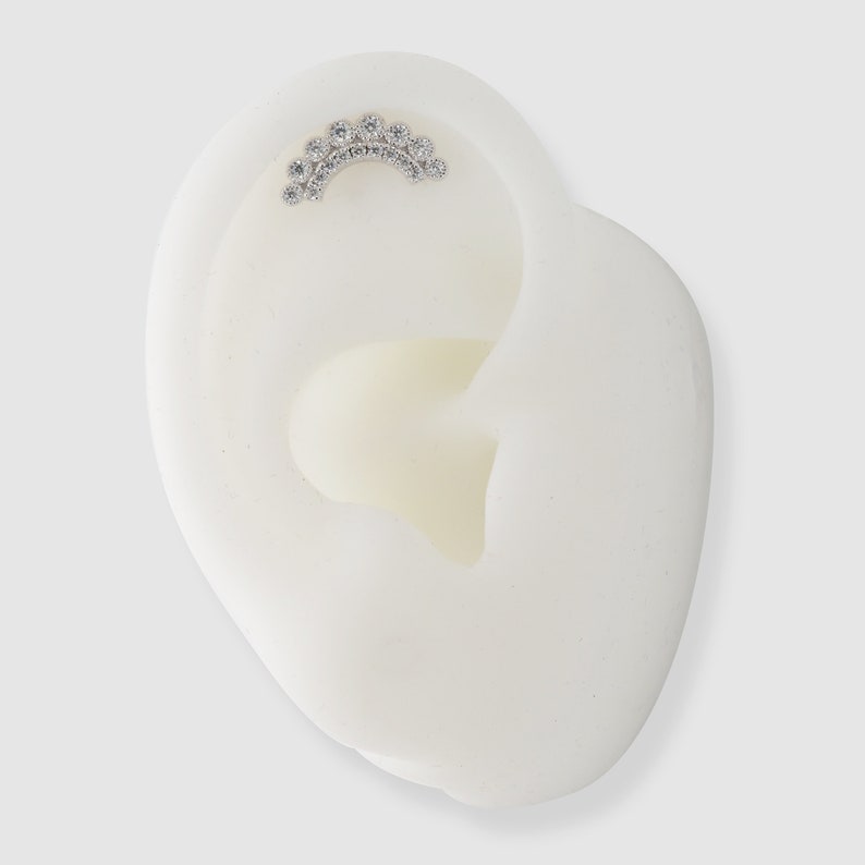 May include: A silver-coloured curved earring featuring several small, clear gemstones. The earring is displayed on a white ear model, highlighting the design and placement of the jewellery. The gemstones are arranged in an arc.