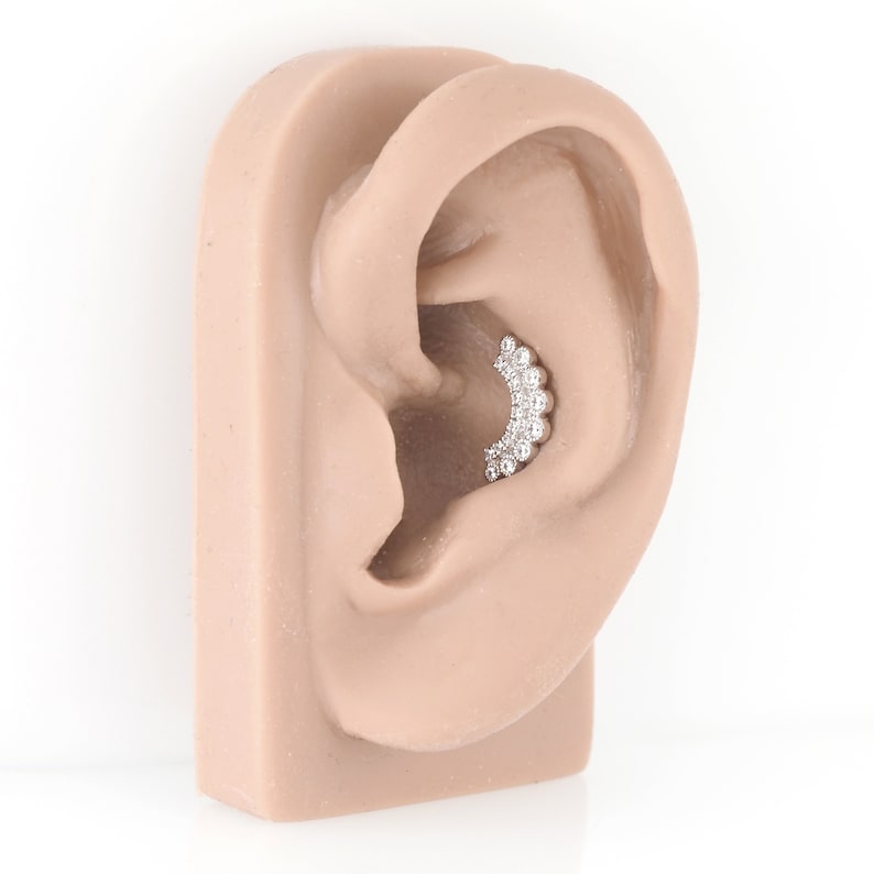 May include: A silver-coloured, curved ear piercing with multiple clear, round gemstones. The jewellery is set in a curved row, designed to fit the curve of the ear. The jewellery is displayed on a light-coloured ear model.