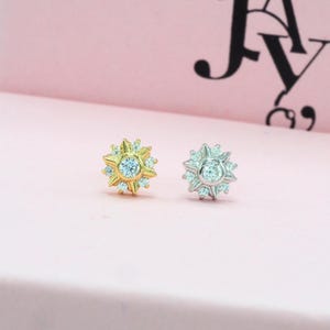 May include: Two starburst-shaped earrings. One is gold-toned, the other is silver-toned. Each earring features a central, round, clear stone surrounded by smaller, clear stones. The earrings are displayed on a light pink surface.