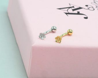 May include: Two belly button rings. One is silver-colored with a flower design and a ball end. The other is gold-colored with a flower design and a ball end. Both are curved and rest on a pink surface.