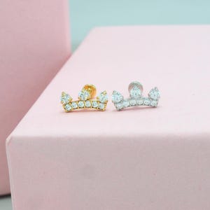May include: Two crown-shaped stud earrings, one gold-toned and one silver-toned, each adorned with multiple clear, sparkling gemstones. The earrings are displayed on a pink surface, with a soft pink background.