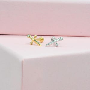 May include: Two small, decorative dagger-shaped earrings. One is gold-colored, and the other is silver-colored. Both earrings have a post and a small, round backing. The earrings are displayed on a pink surface.