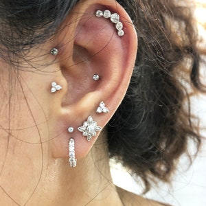 May include: A close-up of a person's ear with multiple piercings. The ear is adorned with a variety of silver earrings, including a hoop earring with small diamonds, a flower-shaped earring with diamonds, a triangle-shaped earring with diamonds, and a curved bar with small diamonds.