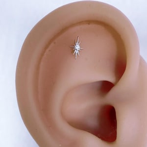 May include: A small, silver-colored ear piercing stud in the shape of a starburst. The stud features a central, round, clear gemstone. The piercing is set on the upper ear cartilage.